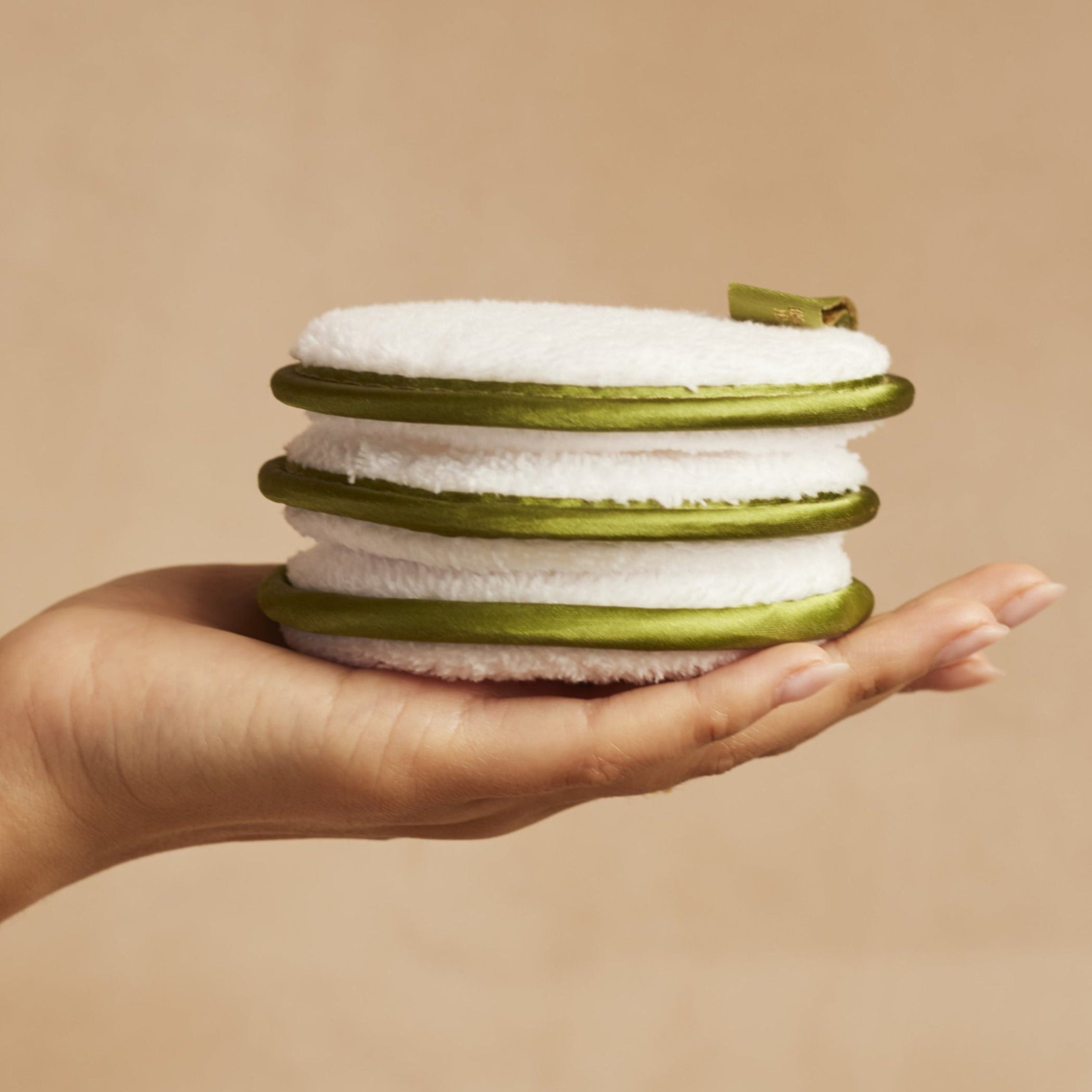 Hand holding a stack of green and white cleansing cloths against a beige background